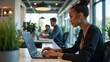 © IgorImageDoc - The image shows a young woman sitting at a desk in an office, working on a laptop. She is wearing a black blazer and has her hair tied up in a bun.