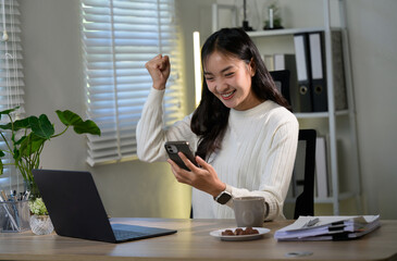 Asian businesswoman is raising her fist in celebration while looking at her smartphone, expressing joy and excitement over a positive outcome in her home office