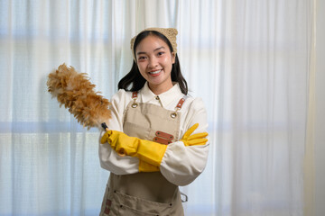 Wall Mural - Young asian housekeeper wearing an apron and yellow gloves, smiling brightly while holding a feather duster, ready to tackle house cleaning tasks with enthusiasm