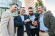 © Jose Calsina - Group of businessmen smiling and discussing something while looking at a cellphone, sharing information or ideas during a work break outdoors, showing teamwork, communication, and collaboration