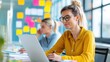 © Vilaysack - A focused woman works on her laptop in a bright office space, surrounded by colorful sticky notes and a colleague in the background.