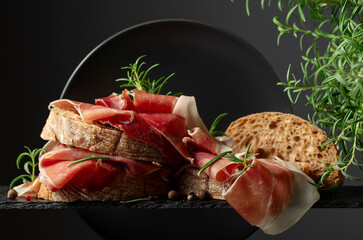 Poster - Ciabatta with prosciutto and rosemary on a black background.