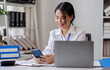 © David - Business asian woman using phone sitting at desk in bright office, working on laptop computer and smiling.
