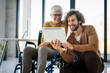 © NDABCREATIVITY - Business woman in wheelchair working with colleague in modern inclusive office. Diversity, support