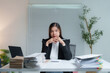 © Tj - Asian businesswoman resting chin on hands at office desk with high piles of paperwork, looking serious and concerned by the amount of work to do