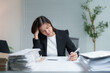 © Tj - Young Asian accountant touching her head and holding pen while calculating financial expense at office desk surrounded by stacks of paperwork