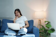 © Tj - Young Asian woman sitting comfortably on a sofa in her living room, engaging in a video call on her laptop, gesturing with her hands while communicating enthusiastically
