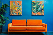 © Pixelite - Un canapé orange vif en velours est placé contre un mur bleu électrique, accompagné de deux tableaux végétaux encadrés et d'une grande plante verte sur le côté gauche.