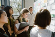 © Maskot - Focused young woman reading adhesive note on bulletin board while standing with students in university