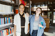 © Maskot - Portrait of smiling young woman standing with hand on shoulder of female friend in university library