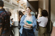 © Maskot - Portrait of smiling young woman holding book while standing near friends in college corridor