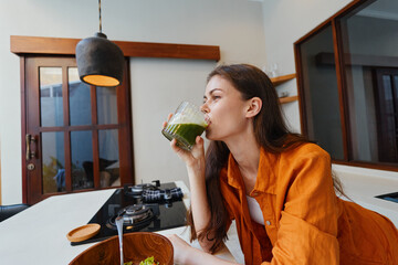 Canvas Print - Young woman enjoying a healthy green smoothie in a modern kitchen, wearing a stylish orange shirt, showcasing a vibrant lifestyle choice and wellness theme