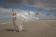 © Milou Dirks - art portrait of woman in long dress standing on beach near ocean waving thin plastic sheet in the wind