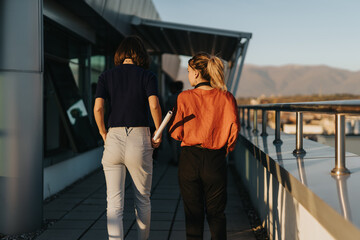 Wall Mural - Two business colleagues walk on an office rooftop, engaging in a discussion about a project. The setting is outdoors with a view of the city, highlighting teamwork and professional collaboration.