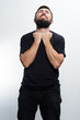 © Niko_Dali - A bearded man in a black shirt and dark pants, against a light background, displays raw emotion, clutching his hands to his chest with his eyes closed and face contorted.