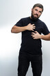 © Niko_Dali - A man with a beard, wearing a black shirt and jeans, scratches his chest with both hands. He appears uncomfortable, suggesting skin irritation or allergy.
