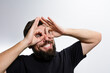 © Niko_Dali - A bearded man with a playful expression forms binoculars around his eyes using his hands, smiling broadly against a light background. His playful gesture conveys a sense of fun and lightheartedness.