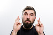 © Niko_Dali - Close-up of a man with a beard, crossing his fingers with a hopeful expression. He's wearing a dark shirt against a white background.