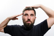 © Niko_Dali - A bearded man in a dark shirt salutes with one hand while adjusting his hair with the other against a white background. His expression is serious.