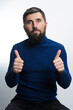 © Niko_Dali - A bearded man wearing a blue turtleneck sweater and dark pants sits against a light background, giving two thumbs up with a positive expression.