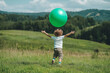 © agnes - A young child with curly hair joyfully playing with a large green ball in a lush open field surrounded by rolling hills and greenery