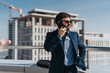 © qunica.com - Confident business professional in a blue suit on a phone call, standing outdoors with a city skyline backdrop. Represents modern business communication and urban corporate lifestyle.