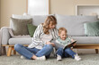 © Pixel-Shot - Happy mother and her little son reading book on floor in living room at home