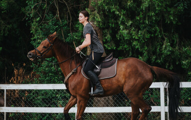 Wall Mural - Young woman confidently riding a horse in an outdoor arena with lush green trees in the background.