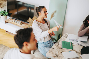 Poster - A group of college students studying together at home, engaging in a casual conversation. The cozy atmosphere and teamwork are evident as they review notes at a marble table.