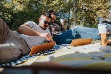 Poster - A young couple sits on a blanket in the park, working on laptops and enjoying coffee under the sunny sky, surrounded by nature's tranquility, symbolizing relaxation and work-life balance.