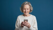 ©  Mohammad Xte - Elderly woman with short wavy hair, wearing a light-colored sweater, smiling as she looks at a smartphone in her hands. The solid blue background highlights her cheerful expression