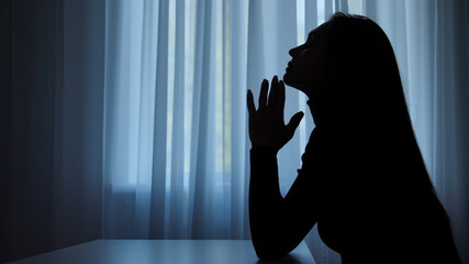 Silhouette of woman suffering mental health issues, sitting against window, depressed expression, touching chin thinking, melancholic indifferent face, side shot.