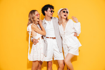 Sticker - Group of young three stylish friends posing in studio. Fashion man and two cute female dressed in casual summer clothes. Smiling models having fun. Cheerful women and guy near yellow wall, sunglasses