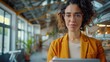 © HARRY - A curly-haired woman wearing glasses and a yellow blazer, standing confidently in a well-lit greenhouse or modern indoor space, radiating intelligence and charm.