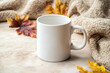 © Nick's Designs - White mug with blank copy space for your advertising text message or promotional content on beige sweater with fall leaves. Porcelain coffee mug mockup on beige stone background