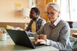© Seventyfour - Medium shot of mature businesswoman wearing elegant glasses typing on laptop keyboard while writing business report sitting at desk in corporate office, copy space