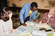 © Seventyfour - Young African American man in denim shirt outfit styled with glasses discussing with female colleagues business strategy pointing at notes on paper, during work meeting at desk in modern office