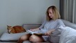 © Alexander - Young adult woman sits comfortably on couch absorbed in book while her cat lounges nearby. In living room caucasian female enjoys book with calm cat resting beside her reading and relaxation at home.