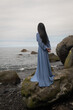 © Milou Dirks - rear view of woman in a long blue dress standing on the beach with rocks and pebbles while looking over the ocean