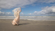 © Milou Dirks - art portrait of woman in long white dress near blue ocean on the beach holding thin fabric on windy day