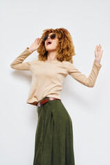 Wall Mural - Fashionable woman expressing joy with curly hair, wearing sunglasses and stylish outfit, beige top and green skirt, standing against a plain light background