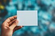 © BerkahStock - Hand holding a blank white card with a blurred background.
