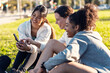 © nenetus - A group of sporty friends women using smartphones after running while sitting on the lawn of the park