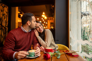 Wall Mural - A happy Caucasian mid-adult couple, with one partner a barista and the other a teacher, enjoys coffee in a cozy cafe. Both are in casual attire, surrounded by warm lighting and a rose on the table.