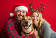 © AlrikaAI - Cheerful middle-aged white couple hugging their dog on red background, man is wearing a Santa hat, woman is wearing reindeer antlers