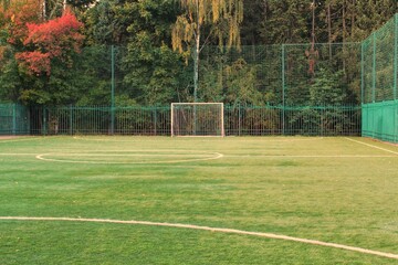 An empty soccer field artificial turf surrounded by green fence and trees. Soccer field surrounded by lush greenery. The turf with goal posts stand tall against a backdrop of vibrant autumn foliage