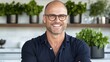 © Leo - Bald middle-aged man with glasses smiling confidently in a plant-filled modern kitchen radiating positivity and warmth in a contemporary lifestyle setting