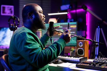 Leinwandbilder - African american composer recording audio files on his song, adding special effects and editing with a mixing console to improve sound quality. Male artist songwriter performing on microphone.
