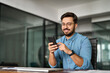 © insta_photos - Busy young Latin business man holding smartphone working in office. Professional happy businessman entrepreneur or manager using financial banking apps making easy payments on cell phone at work.