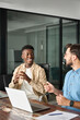 © insta_photos - Vertical photo of two happy busy diverse professional male coworkers software engineers team discussing project together working in teamwork talking sitting at work desk with laptop computers.